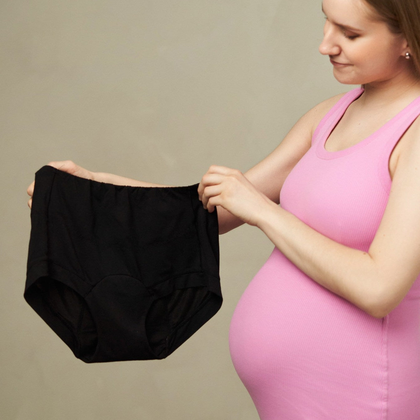 Pregnant woman in a pink dress holding black period underwear against a plain background