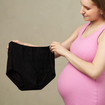 Pregnant woman in a pink dress holding black period underwear against a plain background